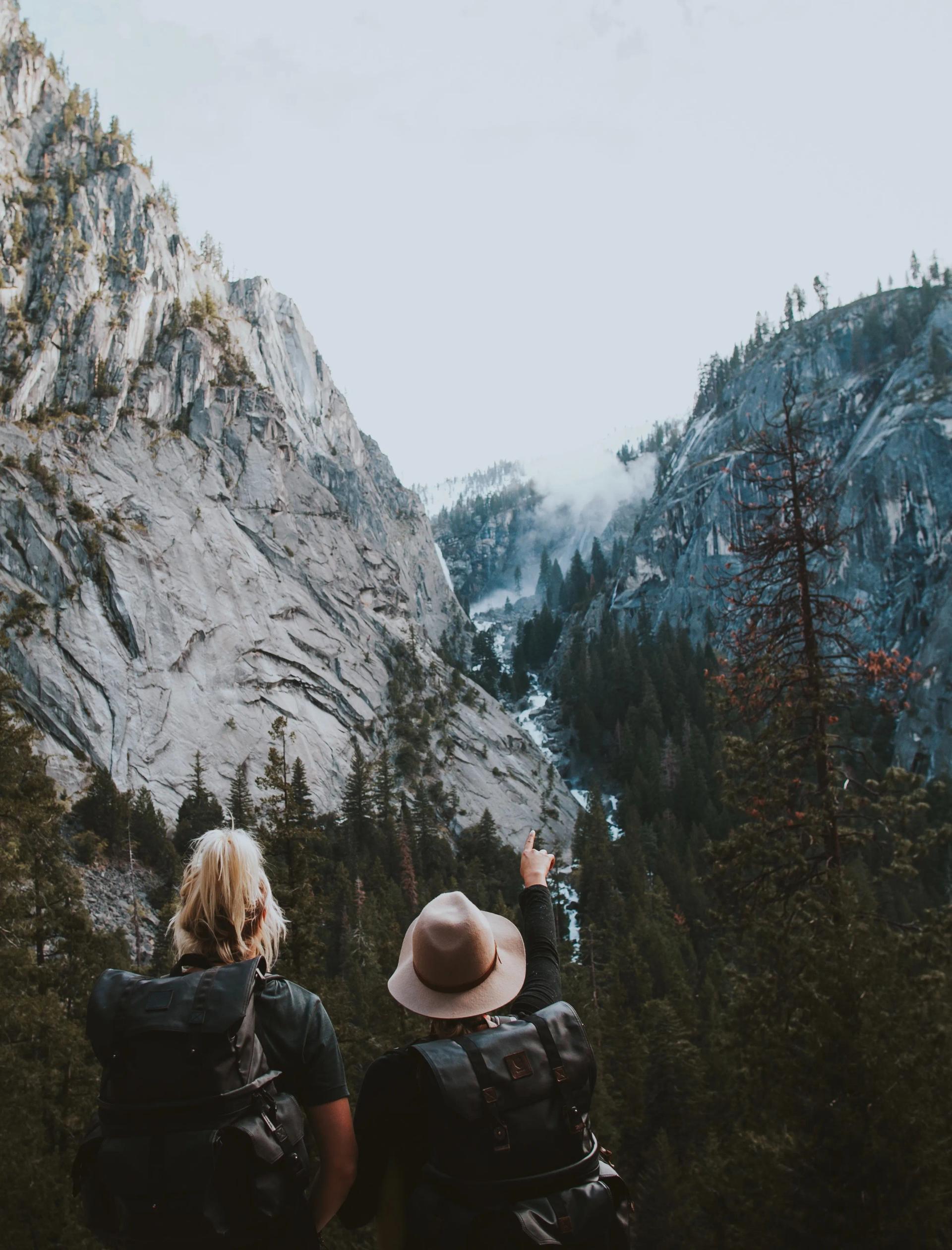 Two hikers on a trail in a mountain range with one pointing at their goal